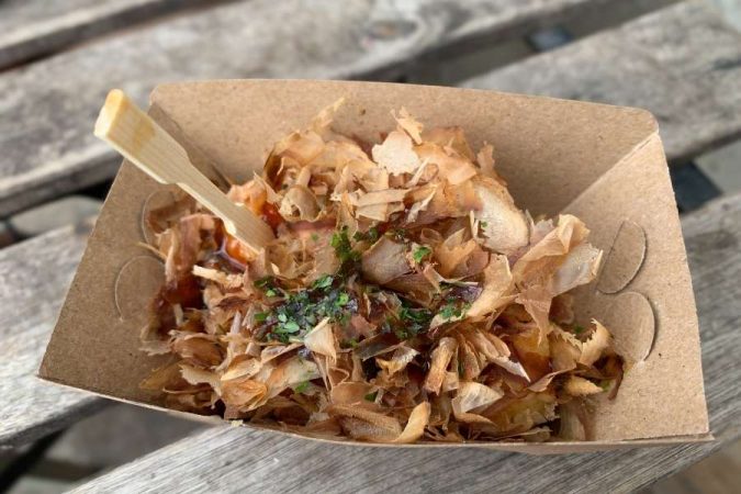Un takoyaki recouvert de flocons de bonite et de sauce, servi dans une barquette en carton kraft avec une fourchette en bois — une nourriture de rue japonaise incontournable.