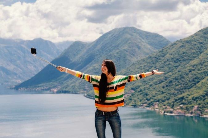 Une femme prend des photos de voyage avec une perche à selfie devant un grand fjord au Monténégro.