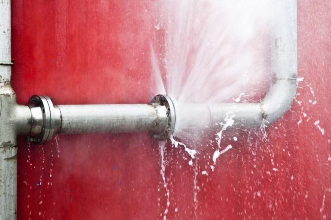 Une importante fuite d'eau jaillit d'une canalisation en métal devant un mur rouge vif, illustrant bien quand les tuyaux fuient.